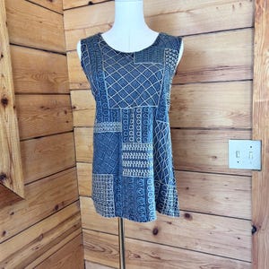 May include: A sleeveless top featuring a dark blue and white patchwork pattern with various geometric shapes, including diamonds, circles, and linear designs. The garment has a round neckline and a straight hem, displayed on a mannequin.