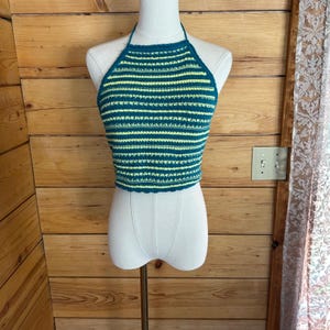 May include: A teal and lime green striped crochet halter top featuring a high neckline and a textured knit pattern. The garment is displayed on a mannequin, showcasing a sleeveless design with horizontal colour bands throughout the bodice.