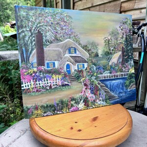 Peut inclure: Une peinture sur toile rectangulaire représentant un cottage au toit de chaume avec une porte bleue et une cheminée, entouré d'un jardin vibrant avec une clôture blanche. Un petit pont traverse un ruisseau bleu sous un ciel doux.