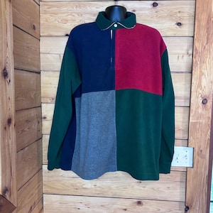 May include: A long-sleeve color-blocked polo shirt featuring a dark green collar and a button-up placket. The garment is constructed with large panels of navy blue, red, dark green, and heather gray fabric arranged in a multi-colored pattern.