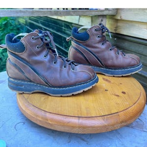 May include: A pair of brown leather hiking boots featuring dark blue padded collars, black laces, and thick dark blue rubber soles. The boots have a rounded toe design with visible stitching along the base and a curved black accent stripe.