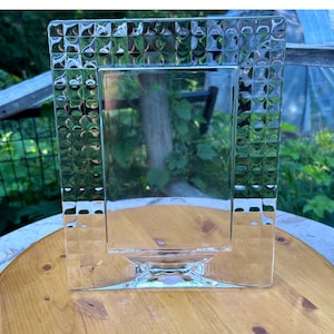 Peut inclure: Un cadre photo rectangulaire en verre transparent. Le cadre présente une large bordure avec un motif en relief en forme de grille composé de petites indentations carrées et alvéolées. Le centre est une vitre rectangulaire lisse et transparente.