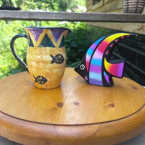 May include: A yellow ceramic pitcher with a dark handle, featuring purple and teal triangular patterns at the rim and black fish silhouettes, alongside a colorful fish-shaped ornament with black, blue, pink, and yellow stripes.