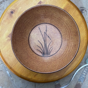 May include: A round ceramic bowl with a speckled tan and brown glaze. The interior features a centred circular design containing a dark brown hand-painted illustration of tall grass or reeds. The bowl has a wide, smooth rim and a deep basin.
