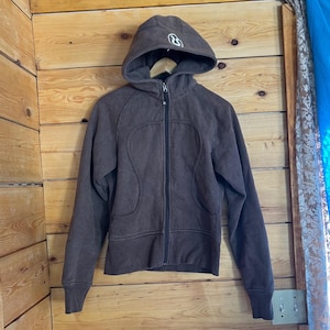 Op de afbeelding: Een bruine hoodie met rits, een zwarte ritssluiting over de hele lengte, lange mouwen en een wit logo geborduurd op de bovenkant van de capuchon. Het kledingstuk heeft twee voorzakken met gebogen naden en geribbelde boorden.