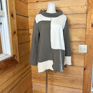 Puede incluir: Una blusa de manga larga con cuello tipo chimenea y un diseño de bloques de color en verde oliva y blanco. La prenda incluye un panel de tela acanalada de color crema en el dobladillo inferior y puños acanalados a juego en las mangas.
