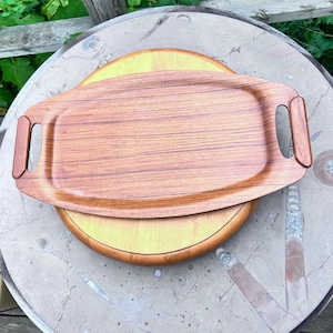 May include: A rectangular wooden serving tray with rounded corners and integrated handles on each side. The tray features a visible brown wood grain pattern throughout its surface, showcasing a smooth, polished finish and a flat, elongated design.