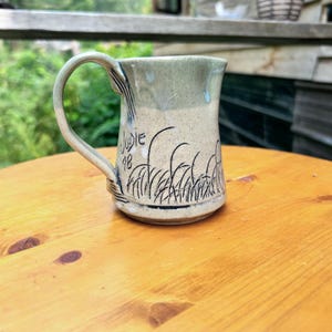Può includere: Una tazza in ceramica con smalto maculato beige chiaro e verde salvia, dotata di un manico curvo e di un disegno a linee nere raffigurante erba alta. Sul lato della tazza è incisa la scritta a mano "JUDIE" sopra il numero "48" in inchiostro nero.