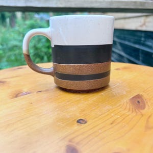 May include: A ceramic mug featuring a speckled off-white upper section, a wide black horizontal band, and a lower section with alternating brown and black stripes. The handle is curved and coloured in a solid brown tone, matching the base.