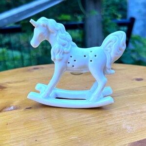 May include: A white ceramic unicorn figurine designed as a rocking horse. The unicorn features a sculpted mane and tail with a series of small holes along its side. It stands on two curved rockers that allow for a gentle back and forth motion.