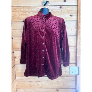 Op de afbeelding: Een bordeauxrode blouse met knopen en een bloemenpatroon. De blouse met lange mouwen is gemaakt van een zacht, fluwelig materiaal en heeft heldere, ronde knopen. De blouse wordt getoond op een zwarte mannequin tegen een houten achtergrond.