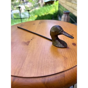 May include: A weathered metal bird head ornament attached to a long, thin, straight metal rod. The bird head features a pointed beak and a dark, tarnished patina with visible signs of age and oxidation across its surface, resting on a flat base.