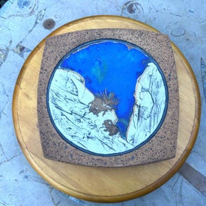 May include: A square ceramic tile with rounded corners featuring a circular center inlay. The center displays a vibrant blue glaze surrounded by textured white clay cliffs and brown organic shapes. The outer border has a speckled brown finish.