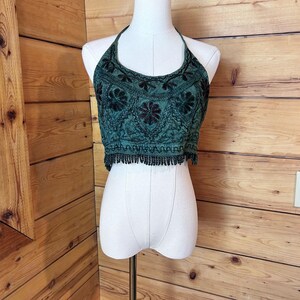 May include: A dark green halter crop top featuring intricate black floral embroidery across the chest and midsection. The garment is finished with a decorative black fringe trim along the bottom hem, creating a textured appearance on the fabric.