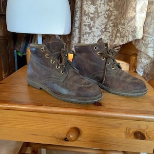 May include: A pair of brown leather ankle boots with laces and brass eyelets. The boots have a worn appearance, suggesting they are well-used. The soles are a darker shade of brown, and the boots are placed on a wooden surface.