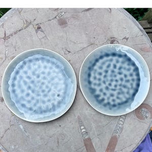 May include: Two round ceramic plates featuring a reactive glaze finish. The left plate displays a light blue and white mottled pattern, while the right plate shows a deeper navy blue and white speckled design with a smooth, glossy surface texture.