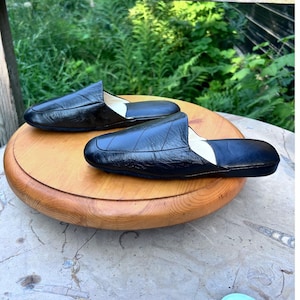 May include: A pair of black leather slip-on mules featuring a smooth, rounded toe design and a low-profile silhouette. The exterior has a subtle textured finish with visible stitching details along the vamp, paired with a light-colored interior lining.