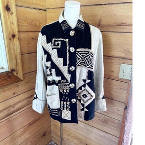 May include: A long-sleeved jacket featuring a black front panel with white embroidered geometric patterns and a beige textured back and sleeves. The jacket has a collared neckline, a front button closure with round buttons, and a patch pocket.