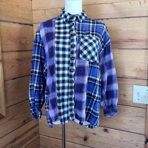 May include: A long-sleeve button-down shirt featuring a patchwork design of blue, black, and white plaid patterns. The shirt has a collar, a single chest pocket on the left side, and a raw, frayed hemline at the bottom, displayed on a mannequin.