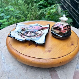 May include: A ceramic set consisting of a square, shallow dish with wavy edges and a small, rounded oil lamp with a white wick. Both pieces feature a glossy glaze with a speckled pattern in shades of pink, black, and white on a cream base.