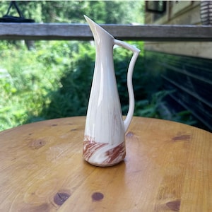 May include: A tall, slender ceramic pitcher with a tapered body and a single curved handle. The surface features a cream-colored base with swirling, marbled patterns in shades of reddish-brown concentrated primarily around the base of the vessel.