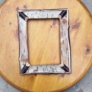 May include: A rectangular picture frame constructed from light-coloured birch bark with a textured, papery surface. The frame features four dark brown, cylindrical wooden accents positioned at each of the four interior corners.