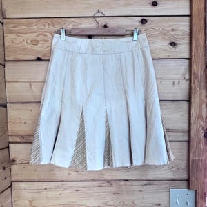 Puede incluir: Falda plisada beige con un panel de acento a rayas, colgada de una percha de madera. La falda está hecha de una tela texturizada y tiene una forma clásica de línea A. El fondo es una pared de madera.