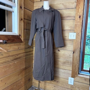 May include: A long, taupe-colored trench coat featuring a classic collar, long sleeves, and a matching fabric belt that ties at the waist. The coat has a button-front closure, a storm flap detail on the chest, and a straight, full-length silhouette.