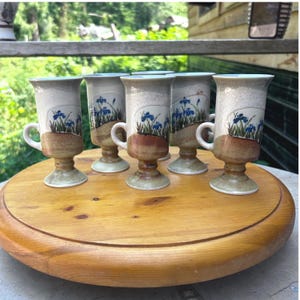 Peut inclure: Un ensemble de cinq tasses en céramique sur pied avec une partie supérieure crème mouchetée et une base émaillée brune. Chaque tasse est ornée d'un motif peint à la main représentant des iris bleus et des tiges vertes sur le corps.