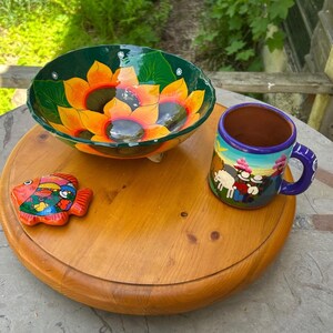 May include: A wooden serving tray displays a hand-painted ceramic bowl with sunflower designs, a colourful mug with a pastoral scene, and a fish-shaped ornament. The bowl is green, orange, and yellow. The mug has a blue handle and rim.