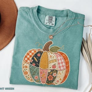 May include: A light green short-sleeve t-shirt featuring a large, central pumpkin graphic composed of various patchwork-style patterns including plaid, floral, apple, pencil, and alphabet motifs. The shirt has a Comfort Colors brand label.