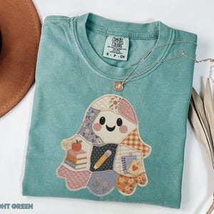 May include: A light green t-shirt featuring a central ghost-shaped graphic composed of various patterned fabric patches. The ghost design includes illustrations of a stack of books, an apple, a pencil, and a notebook. The shirt has a Comfort Colors label.