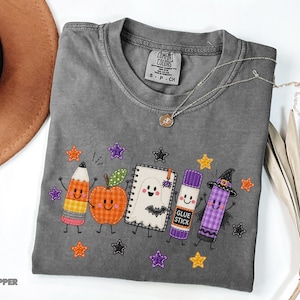 May include: A gray short-sleeved t-shirt featuring a graphic of a smiling pencil, apple, ghost-covered notebook, glue stick, and witch-hat-wearing crayon. The design includes small orange, purple, and black stars on a Comfort Colors size S-CH label.