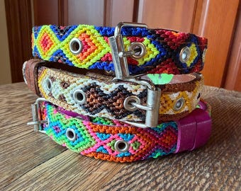 May include: Three colourful dog collars stacked on a wooden surface. The collars feature woven patterns in vibrant colours like blue, yellow, red, and pink. Each collar has a metal buckle and eyelets.