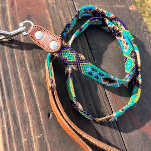 May include: A patterned dog lead in teal, purple, green, and brown. It features a woven design and a brown leather handle. A silver metal clasp is attached. The lead rests on a wooden surface.