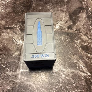 May include: A grey rectangular ammunition box featuring a blue bullet design and the text ".308 WIN". The box is made of a textured material.