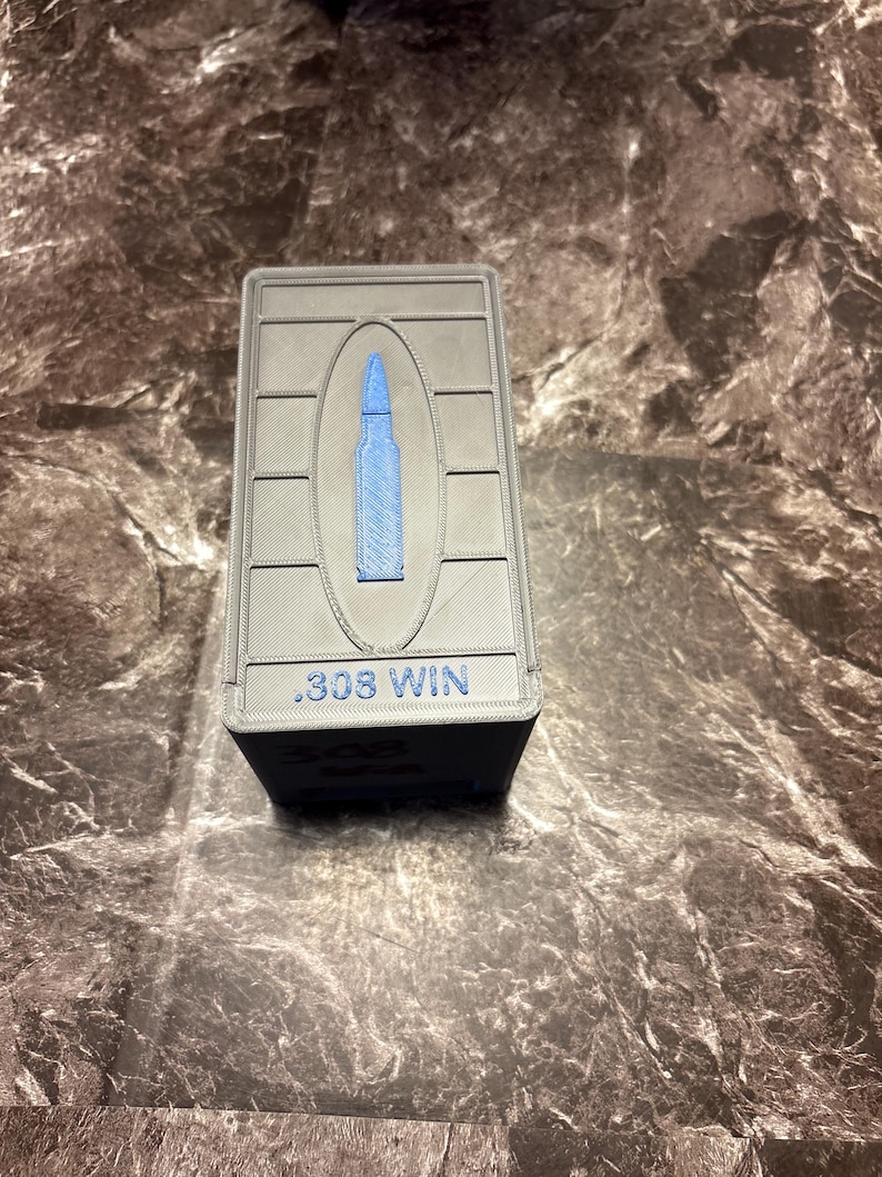 Puede incluir: Una caja de munici&oacute;n rectangular gris con un dise&ntilde;o de bala azul y el texto ".308 WIN". La caja est&aacute; hecha de un material texturizado.