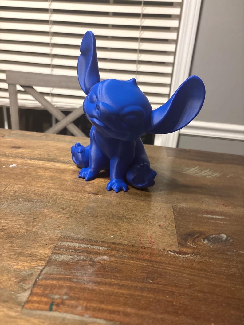 May include: A blue figurine of a cartoon character with large ears, sitting on a wooden table. The figurine is a solid blue colour and has a detailed design. The background includes a window with blinds and a chair.