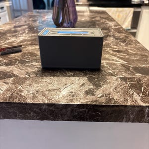 Puede incluir: Un dispensador de pa&ntilde;uelos rectangular gris con un detalle azul en la tapa. Est&aacute; colocado sobre una encimera de m&aacute;rmol marr&oacute;n y gris. Un jarr&oacute;n morado es visible en el fondo. La encimera mide aproximadamente 100 cm de ancho.