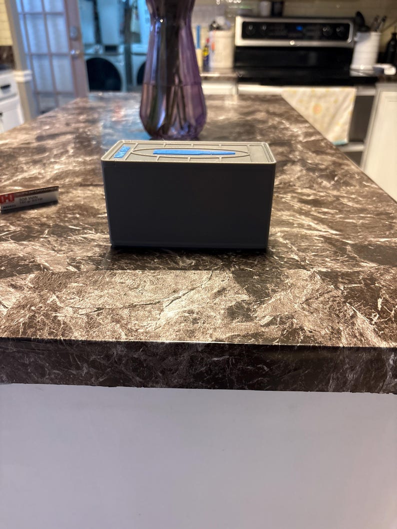 Puede incluir: Un dispensador de pa&ntilde;uelos rectangular gris con un detalle azul en la tapa. Est&aacute; colocado sobre una encimera de m&aacute;rmol marr&oacute;n y gris. Un jarr&oacute;n morado es visible en el fondo. La encimera mide aproximadamente 100 cm de ancho.