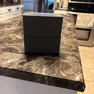Puede incluir: Una caja rectangular de dos piezas, gris oscuro, con un acento azul en la parte superior. La caja est&aacute; sobre una encimera con un patr&oacute;n de m&aacute;rmol marr&oacute;n y gris. La encimera es parte de una isla de cocina blanca.