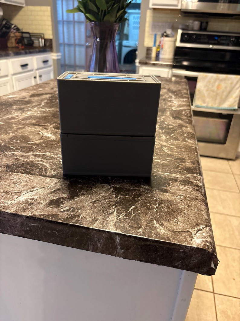Puede incluir: Una caja rectangular de dos piezas, gris oscuro, con un acento azul en la parte superior. La caja est&aacute; sobre una encimera con un patr&oacute;n de m&aacute;rmol marr&oacute;n y gris. La encimera es parte de una isla de cocina blanca.