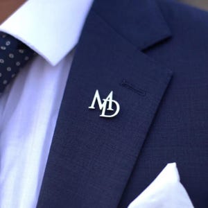 May include: A silver lapel pin with the intertwined letters "M" and "D" is affixed to a navy blue suit jacket. The jacket is paired with a white dress shirt and a patterned navy blue tie. A white pocket square is visible.