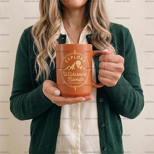 May include: A large, orange ceramic mug with the text "EXPLORE Wilderness Blends PREMIUM ROASTS" in a compass-style design. The mug has a handle and is held by a person wearing a green cardigan and a white shirt.