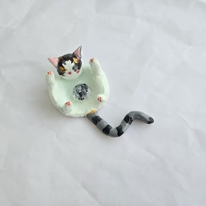 May include: A whimsical handmade cat figurine. The cat is lying on its back with its paws up, and has a black, white, and tan face. The figurine has a gray and black striped tail. The cat is resting on a light green base with a dark gray ball.