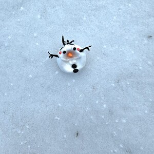 May include: A small, round snowman figurine with a white body, black button eyes, and a carrot nose. It has black twig arms and a twig hair. The snowman is set against a snowy background.