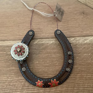 May include: A decorative horseshoe ornament with a brown finish, adorned with red and gold floral accents. The horseshoe features a circular embellishment with a red floral design and turquoise accents. Small gold-colored studs and a hanging string complete the design.