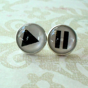 Op de afbeelding: Een paar zilveren oorbellen met zwarte en witte symbolen. De linker oorbel heeft een zwarte driehoek die naar rechts wijst. De rechter oorbel heeft twee verticale zwarte balken.