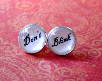 Puede incluir: Un par de pendientes de plata con cabujones redondos blancos. El pendiente izquierdo dice "Don't" y el pendiente derecho dice "Blink".