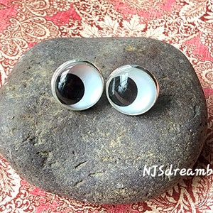 May include: Two large, white googly eyes with black pupils, set against a gray rock. The eyes are silver-rimmed and have a glossy finish.